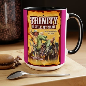 Puede incluir: Una taza de cerámica blanca con asa y borde negros. La taza presenta una ilustración de estilo vintage de un hombre a caballo con el texto "TRINITY IS STILL MY NAME" y "Those Trinity Boys Are Back!" en una pancarta amarilla. El texto "TERENCE HILL & BUD SPENCER" también es visible en la imagen.