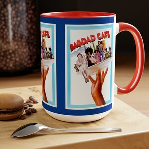May include: A red and white ceramic mug with a blue and white design featuring the text "Bagdad Cafe". The mug has a red handle.
