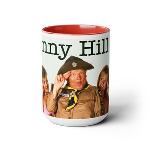 May include: White ceramic mug with a red rim. The mug features a photo of a man in a scout uniform with two women. The text "Benny Hill" is printed in black at the top of the mug.