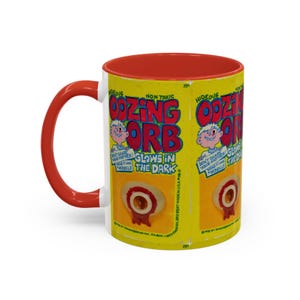 Könnte beinhalten: Eine weiße Keramiktasse mit rotem Rand und Henkel. Die Tasse zeigt eine gelbe und rote Grafik eines Cartoon-Auges mit dem Text "Oozing Orb" und "Glows in the Dark".
