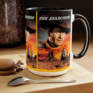 Può includere: Tazza in ceramica bianca con un design del film "The Searchers". Il design presenta un ritratto di un uomo con cappello da cowboy su uno sfondo di tramonto nel deserto. La tazza ha un manico e un bordo neri.