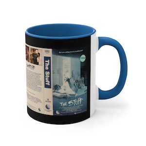 Puede incluir: Una taza de café de cerámica blanca con un borde y un asa azules. La taza presenta una imagen en blanco y negro de una cinta VHS para la película de terror de 1985 "The Stuff". El texto en la cinta VHS dice "Are you eating it or is it eating you?" y "The Stuff".