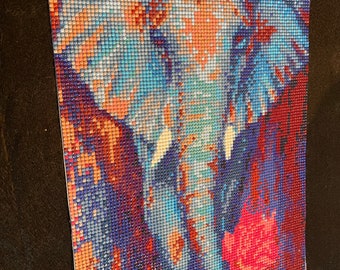 May include: A close-up of a diamond painting depicting an elephant with blue and red tones. The elephant is facing the right side of the image and has a pink flower in its trunk.