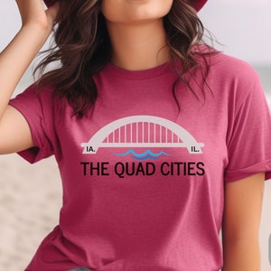 May include: A heather raspberry t-shirt with a white graphic of a bridge over water with the text "THE QUAD CITIES" and "IA." and "IL." on either side of the bridge.