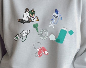 May include: A gray sweatshirt with a graphic print of medical equipment, including a stethoscope, an oxygen mask, a nebulizer, and a medical device with a tube. Two people are shown performing CPR on a person lying on the ground.
