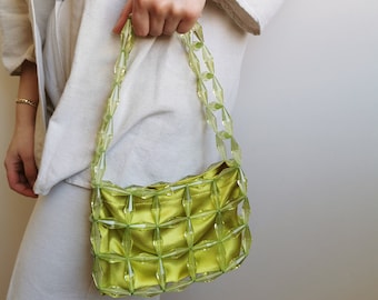 May include: A lime green handbag with a grid-like design made of clear, faceted beads. The bag's interior is a matching satin fabric. The bag is held by a person wearing a cream-colored linen jacket and pants.