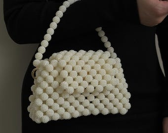 Pode incluir: Uma bolsa branca de contas com um design tecido. A bolsa tem uma alça curta e um fecho dourado.