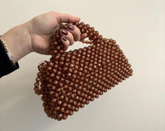 Puede incluir: Un bolso de mano de cuentas marrones con asa superior. El bolso está hecho de pequeñas cuentas redondas que están entrelazadas.