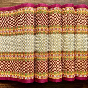 May include: A rolled-up, rectangular woven placemat with a vibrant pattern. The design features alternating bands of red, yellow, green, and white, with a deep pink border. The intricate weave creates a textured surface.