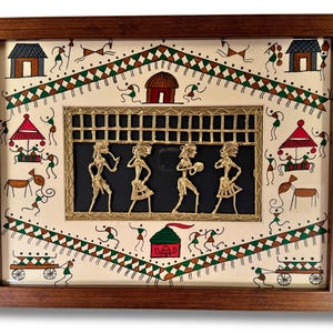May include: A framed artwork featuring a traditional Indian folk art scene with a central panel of four figures in gold metalwork. The figures are dancing and playing instruments. The background is painted with a white background and colourful geometric patterns.