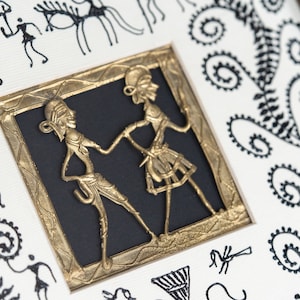 May include: A framed artwork featuring a gold metal cutout of two figures dancing. The figures are stylized and have a tribal aesthetic. The artwork is surrounded by a black mat and a gold frame. The background of the artwork is white with black tribal designs.