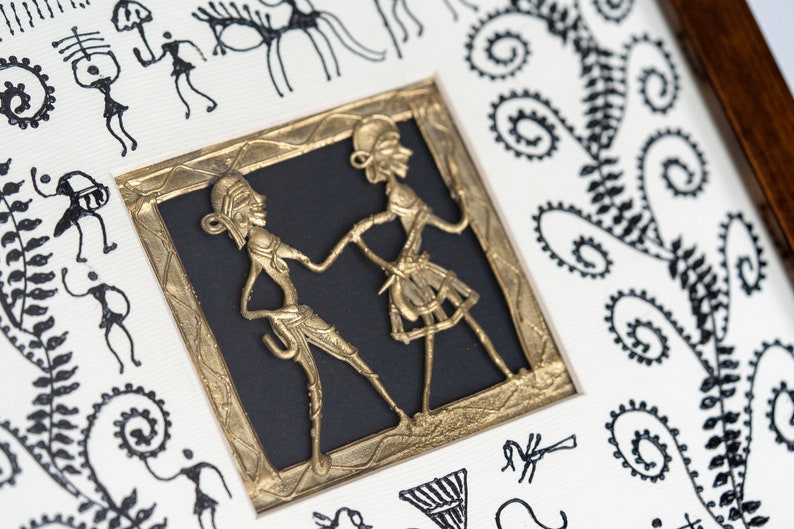 May include: A framed artwork featuring a gold metal cutout of two figures dancing. The figures are stylized and have a tribal aesthetic. The artwork is surrounded by a black mat and a gold frame. The background of the artwork is white with black tribal designs.