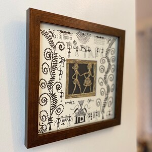 May include: A framed artwork with a white background and black line drawings of people, animals, and plants. The artwork is surrounded by a dark brown frame.