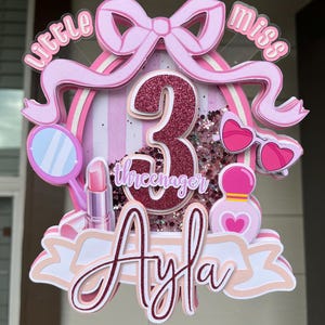 May include: A pink and white birthday cake topper with the words "little miss" and "Ayla" displayed. The number "3" is in the center, surrounded by glitter and decorative elements like sunglasses and lipstick.