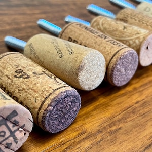 May include: A close-up of a collection of cork bottle stoppers with various labels and designs. The stoppers are made of natural cork and have a variety of colors and textures. The stoppers are arranged in a row, with the labels facing the camera. The labels include the text "Fondee en 1859", "Stouwers", "D.D. Editions", "Diavlo", and other text.