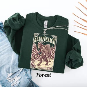 May include: A dark forest green crewneck sweatshirt featuring a graphic print of Krampus in a snowy forest setting. The text reads KRAMPUSNACHT and 5. DEZEMBER. The design includes a lantern, a basket, and bells. The sweatshirt is paired with blue jeans.