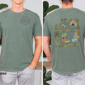 Pode incluir: T-shirt verde musgo com o texto "TIRED DAD CLUB" na frente. A parte de trás apresenta um gráfico colorido com tema de dinossauros com um design de mapa rodoviário, dinossauros e um vulcão. A camisa é feita de um material macio.