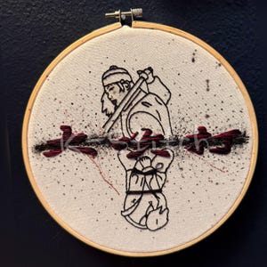 May include: Embroidered artwork in a wooden hoop. The design features a black and red illustration of a figure with a staff, and Japanese text. The background is white with red and black speckles.