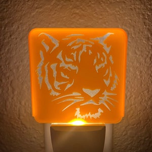May include: Orange night light with a white tiger silhouette. The night light plugs into a standard electrical outlet.