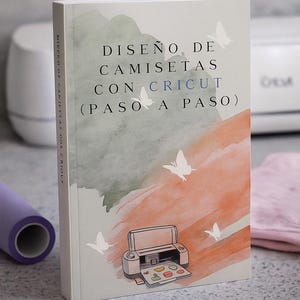 Puede incluir: Un libro titulado "DISEÑO DE CAMISETAS CON CRICUT (PASO A PASO)" presenta un diseño de acuarela con mariposas y una ilustración de máquina Cricut. La portada tiene una paleta de colores suaves de naranja, verde y blanco. Se ve un rollo de vinilo morado y un paño rosa.