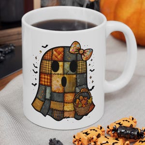 Op de afbeelding: Witte keramische mok met een Halloween spookontwerp. Het spook is gemaakt van patchwork vierkanten in oranje, bruin en grijs, met een strik en een mandje met bladeren. Zwarte vleermuizen en sterren omringen het spook.