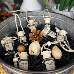 May include: A collection of six small glass bottles with wooden tops and string hangers. The bottles are filled with a clear liquid and have labels that read "Country Road Candle Co." and various scents, including "Angel Wings", "Fresh Linen", and "Mahogany". The bottles are arranged in a metal bucket with black shredded paper and decorative items.