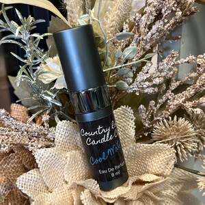 Puede incluir: Una peque&ntilde;a botella negra con tapa plateada y una etiqueta blanca que dice "Country Candle Co. Cool Water Eau De Parfum 8 ml".