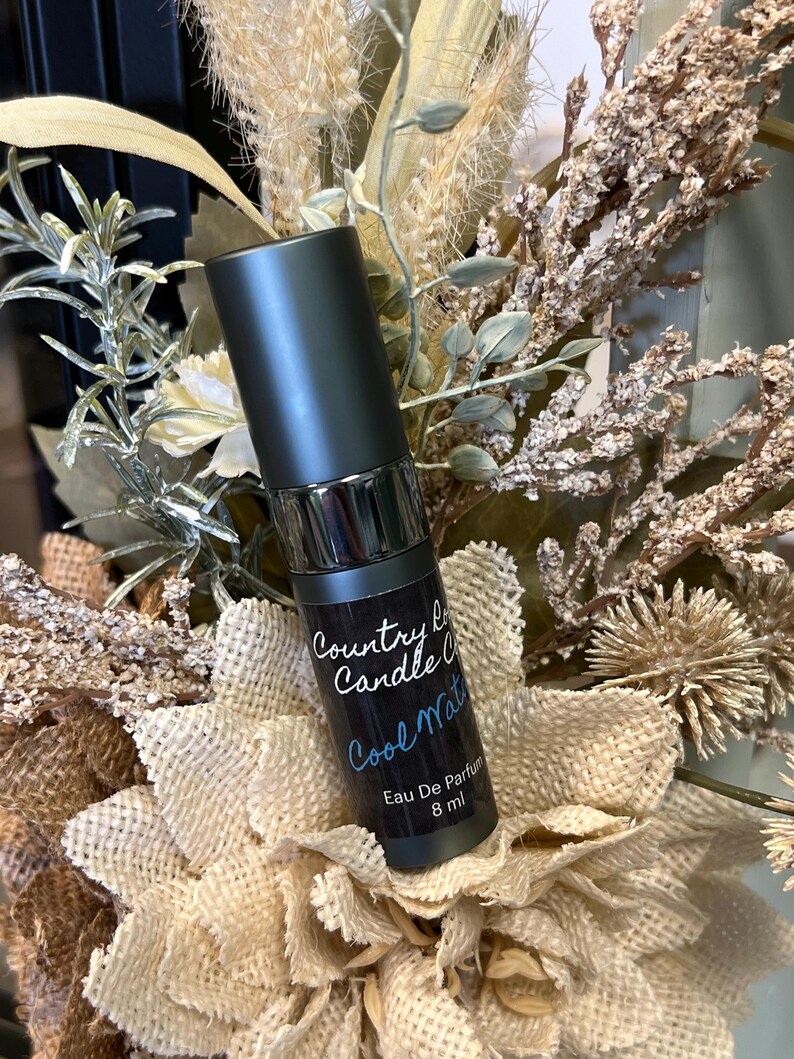 Puede incluir: Una peque&ntilde;a botella negra con tapa plateada y una etiqueta blanca que dice "Country Candle Co. Cool Water Eau De Parfum 8 ml".