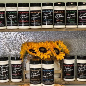 May include: A shelf display of white plastic jars with black lids. The jars are labeled "Carpet Refresher" with various scents, including "Lodge", "Laundry Day", "Apple Easy", "Man Cave", "Volcano", "Mahogany", "Lavender & Chamomile", "Bum Bum", "Spring Flower", "Sandalwood", "Fresh Linen", "Love", and "Bear Claw".