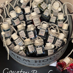 May include: A metal bucket filled with small glass bottles with cork tops and string hangers. The bottles are filled with a clear liquid and labeled with the text "Country Road Candle Co." and various scents, including "Cherry", "Driftwood", "French Vanilla", "Angel Wings", and "Twilight".