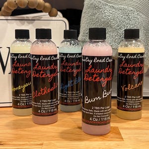 May include: Five bottles of laundry detergent with black caps and labels. The labels read "Country Road Candle" and feature names like "Honeyou" and "Volcano." Each bottle contains 4 fl oz / 118 ml of liquid.