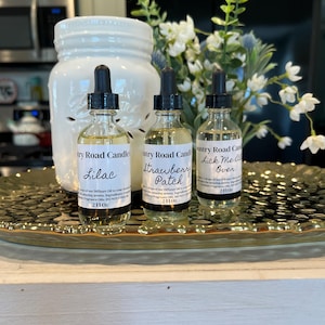 May include: Three glass bottles of diffuser oil with black droppers, labeled "Lilac", "Strawberry Patch", and "Lick Me All Over". The bottles are on a decorative tray with a white jar in the background. The bottles are 2 fl oz each.