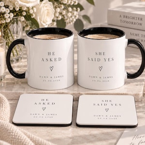 May include: Two white ceramic mugs with black handles and rims, each featuring the text HE ASKED or SHE SAID YES, a heart icon, and the names DAWN & JAMES with the date 20.09.2026. Two matching square white coasters display the same printed text and design.