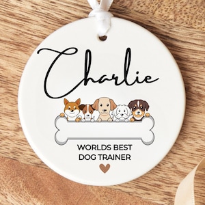 May include: White ceramic ornament with the name "Charlie" written in black cursive. The ornament features a bone with five cartoon dogs peeking over the top. The bone has the text "Worlds Best Dog Trainer" and a small brown heart.
