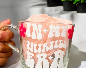 May include: A clear glass mug with a white design that says "In My Thirties Era" in a retro font. The design also includes pink flowers.