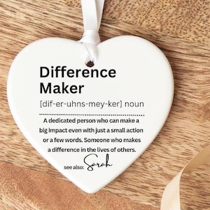 May include: A white heart-shaped ornament with the text "Difference Maker" and its definition. The ornament hangs from a white ribbon and rests on a wooden tray. A cream-coloured ribbon is also present.