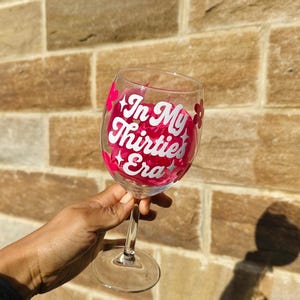 May include: A clear wine glass with a long stem, featuring the text "In My Thirties Era" in white script with pink accents. The glass is held in front of a brick wall.