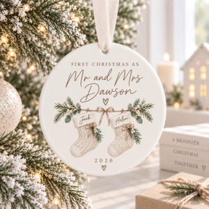 Puede incluir: Un adorno redondo de cerámica blanca que cuelga de una cinta color crema. Presenta el texto "FIRST CHRISTMAS AS Mr and Mrs Dawson" sobre una ilustración de dos calcetines etiquetados "Jack" y "Helen" con ramas de pino y el año "2026".