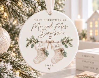 Könnte beinhalten: Ein rundes, weißes Keramikornament, das an einem cremefarbenen Band hängt. Es zeigt den Text "FIRST CHRISTMAS AS Mr and Mrs Dawson" über einer Illustration von zwei Strümpfen mit den Namen "Jack" und "Helen", Tannenzweigen und dem Jahr "2026".