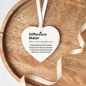 Heart shaped ceramic ornament. With a definition of a difference maker on it , in black text. The picture shows the heart on a wooden tray with a ribbon beside it.
