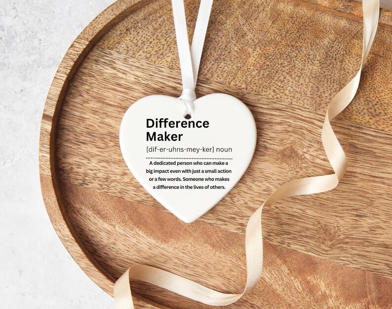 Heart shaped ceramic ornament. With a definition of a difference maker on it , in black text. The picture shows the heart on a wooden tray with a ribbon beside it.