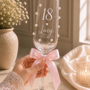 May include: A clear glass champagne flute featuring the number 18 and the name Lucy printed in white, accented with small white pearls along the sides and a light pink satin ribbon tied in a bow around the stem of the glass.