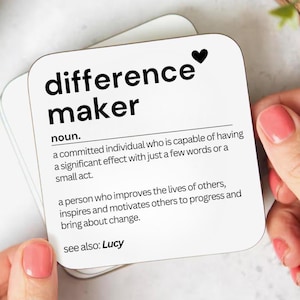 May include: White coaster with black text that reads "difference maker" and a definition of the word. The definition reads "a committed individual who is capable of having a significant effect with just a few words or a small act. a person who improves the lives of others, inspires and motivates others to progress and bring about change. see also: Lucy"