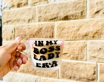 May include: A clear glass mug with a handle, filled with a creamy beverage. The mug features the text "IN MY BOSS LADY ERA" in black lettering. Pink floral accents are on the top of the mug. The mug is held in front of a textured brick wall.