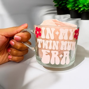 Puede incluir: Taza de vidrio transparente con las palabras "IN MY TWIN MUM ERA" en una fuente retro. El texto es en blanco y rosa, con detalles de flores rojas. La taza est&aacute; llena de una tela rosa claro.