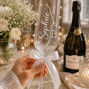 May include: A clear glass champagne flute featuring the white printed text Sophie and 30th, decorated with small white pearls along the rim and a white satin ribbon tied in a bow around the stem of the glass.