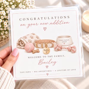 Puede incluir: Una tarjeta de felicitación cuadrada con el texto CONGRATULATIONS on your new addition, WELCOME TO THE FAMILY, Bailey, TINY PAWS BIG ADVENTURES A LIFETIME OF LOVE. Las ilustraciones incluyen una manta con huellas, una pelota, un collar y un cuenco.
