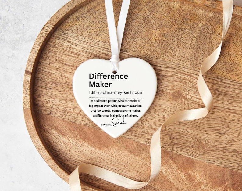 Heart shaped ceramic ornament. With a definition of a difference maker on it , in black text. The ornament comes personalised with a name. The picture shows the heart on a wooden tray with a ribbon beside it.