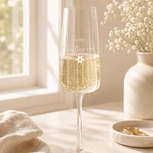 Puede incluir: Una copa de champán de cristal transparente llena de un líquido espumoso. El cristal presenta el texto impreso en blanco "in my thirties era" sobre un pequeño gráfico de una margarita blanca y amarilla. El tallo es fino y la base es circular.