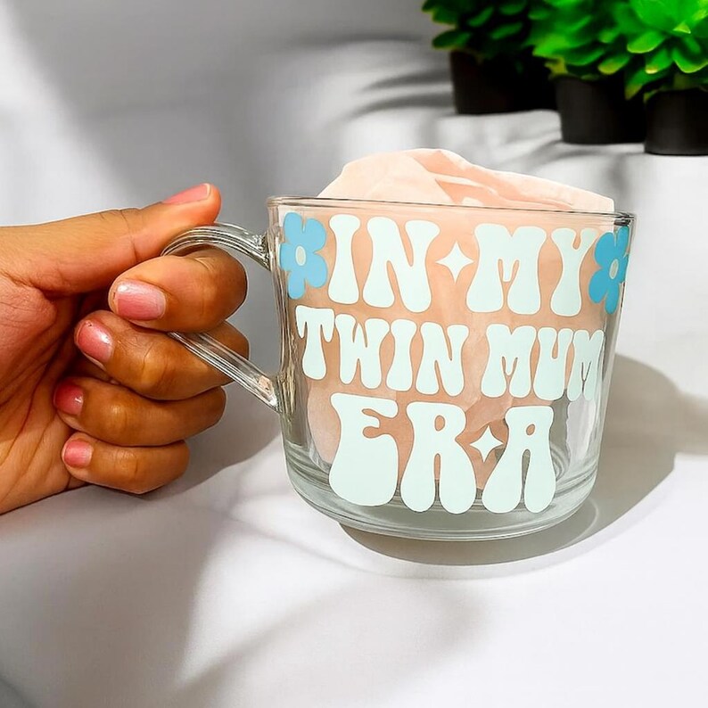Puede incluir: Taza de vidrio transparente con asa, con el texto "IN MY TWIN MUM ERA" en blanco y azul claro. El dise&ntilde;o incluye detalles florales en azul claro. La taza est&aacute; rellena con un material rosa claro.